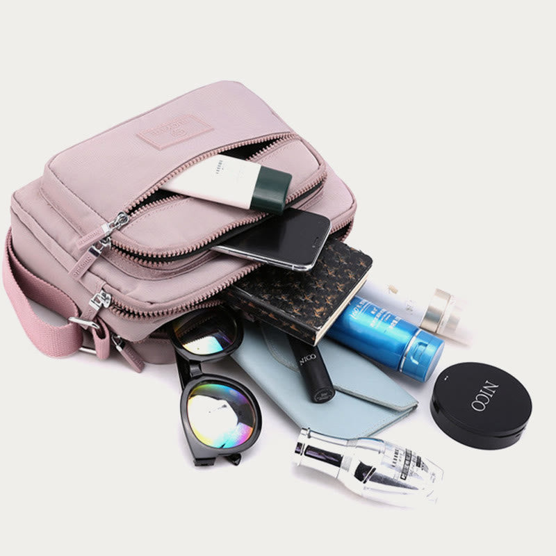 Light pink four-zip crossbody bag shown with phone, wallet, bottle, and small essentials
